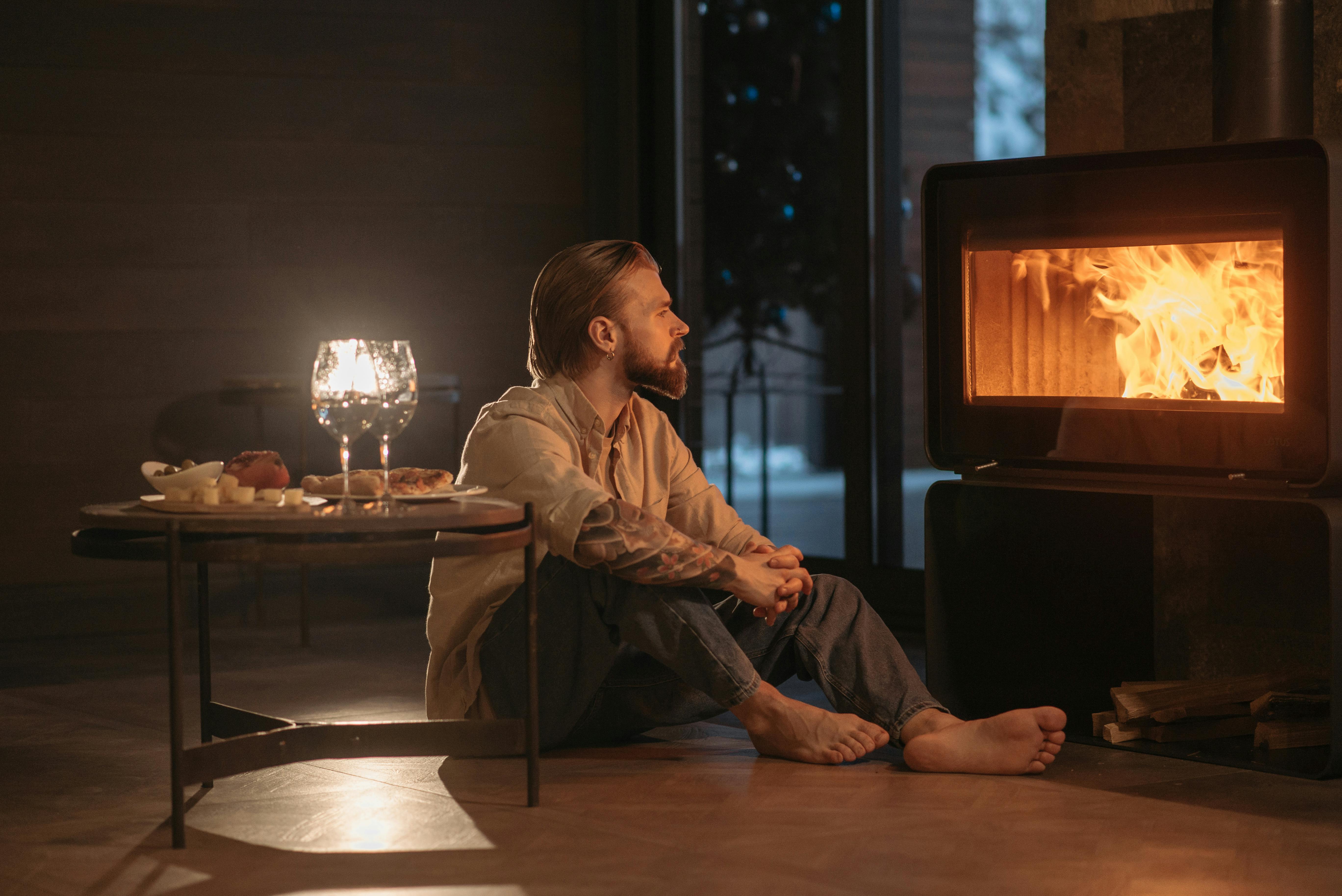 A man with tattoos relaxes by a warm fireplace, exuding a cozy atmosphere indoors.