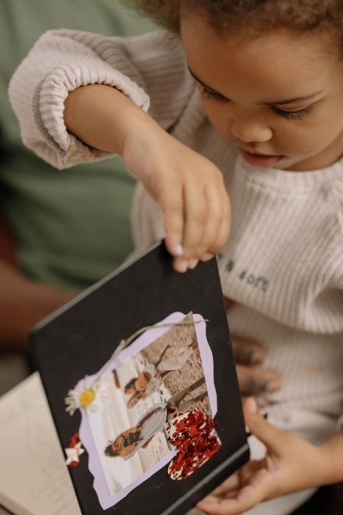 Free A young child curiously examines a family photo album indoors, depicting a moment of exploration and nostalgia. Stock Photo