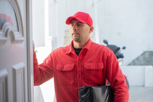 A Delivery Man in Red Long Sleeves and Cap Holding a Parcel while Standing Beside the Door