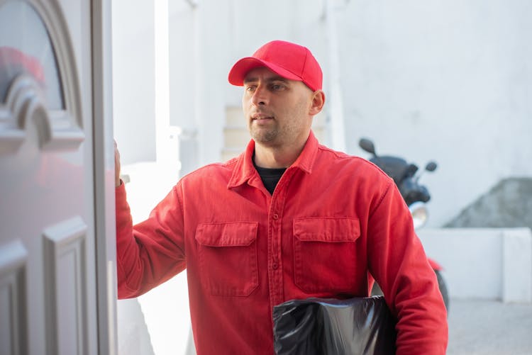 A Delivery Man In Red Long Sleeves And Cap Holding A Parcel While Standing Beside The Door