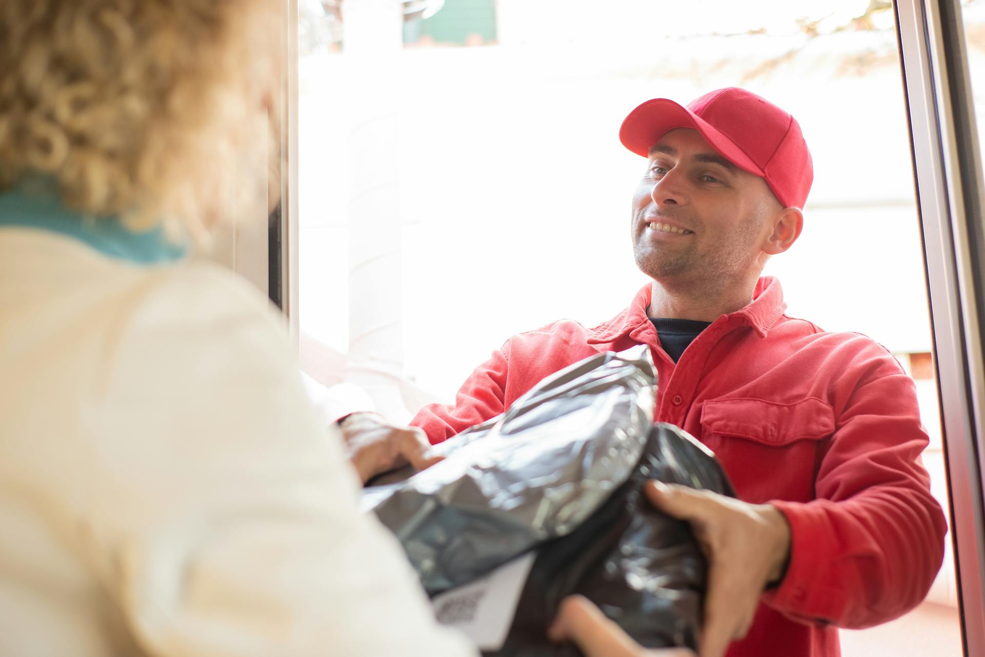 A smiling delivery man in red uniform hands over a package to a customer by the door.