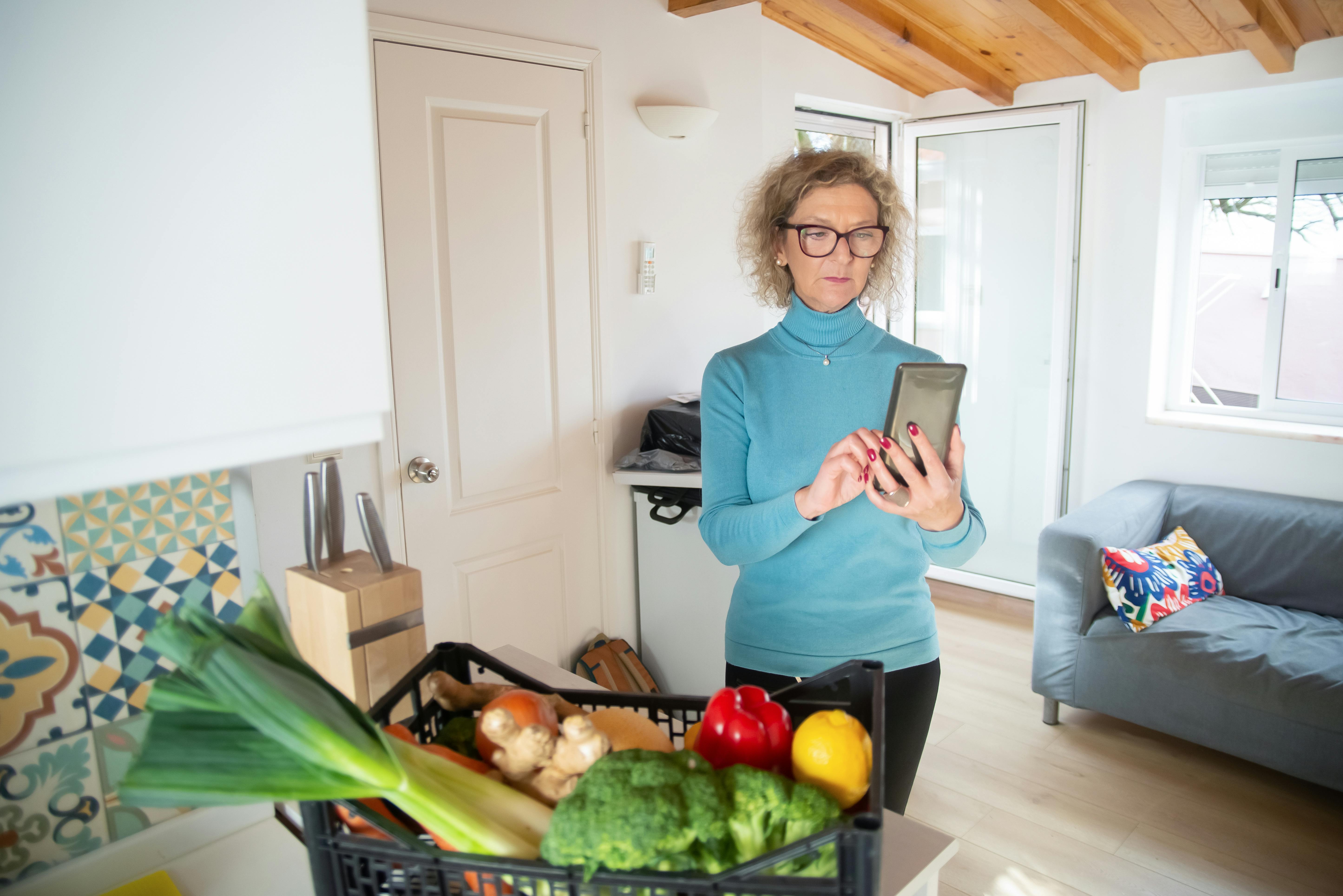 Elderly woman checks her smartphone in a cozy kitchen with fresh vegetables.