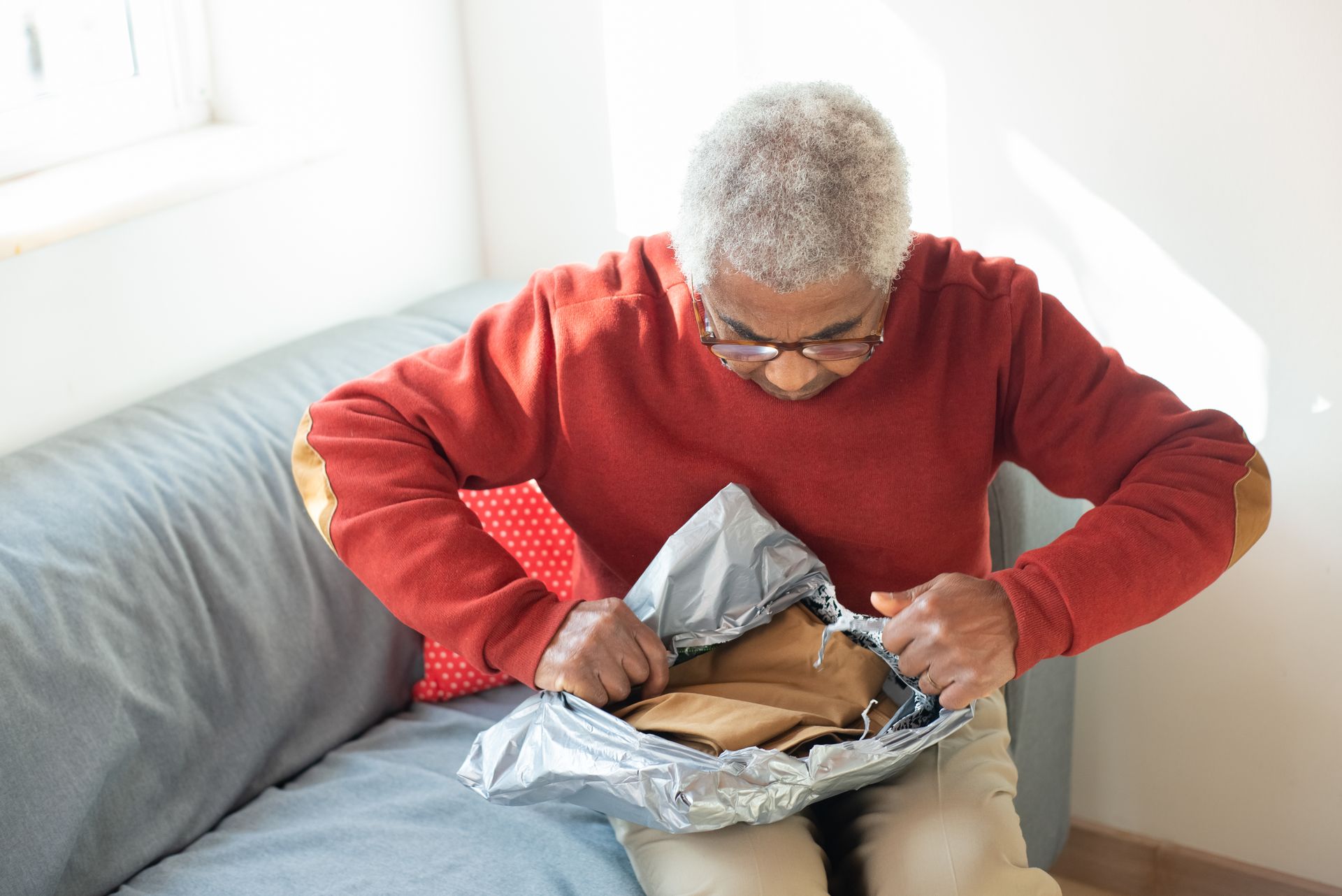 A senior man in a red sweater opens a package on a sofa indoors.