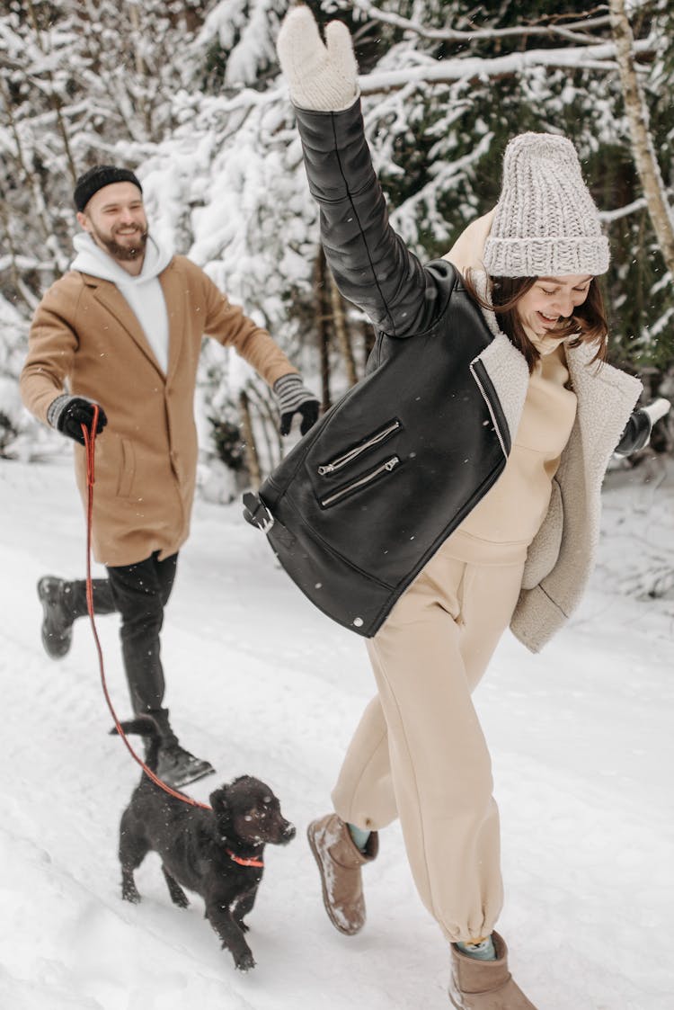 Smiling Woman With Arm Raised And Man Walking Dog In Forest In Winter