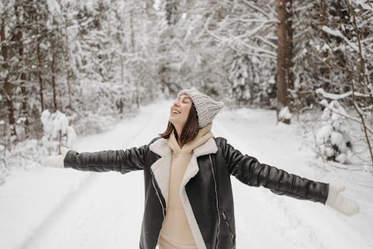 Woman in winter clothing embracing snowfall joyfully in a snowy forest.