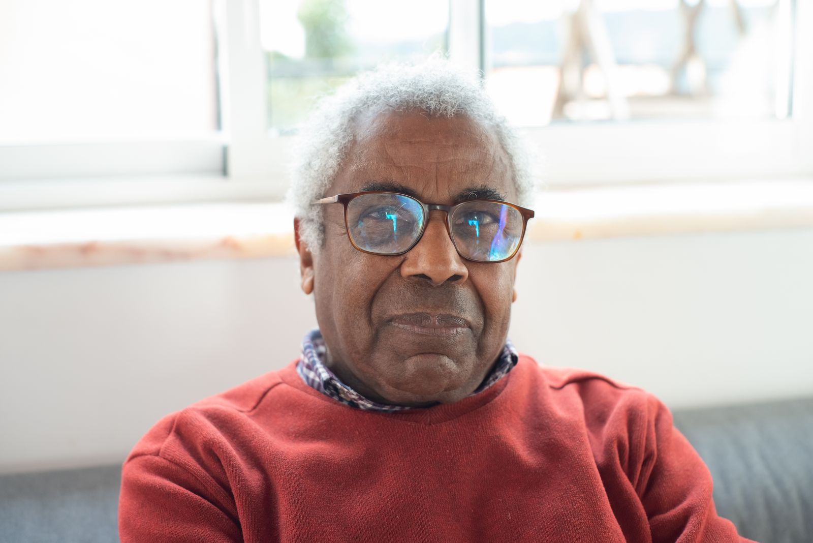 An older Black man with gray hair and glasses seated indoors, looking off to the side, in the age group most affected by rheumatoid arthritis and most often diagnosed late.