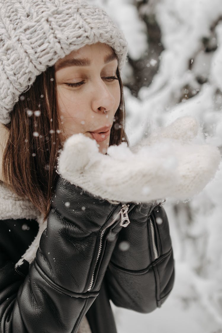 Woman With A Gray Knitted Cap Blowing White Snow On Her Hands