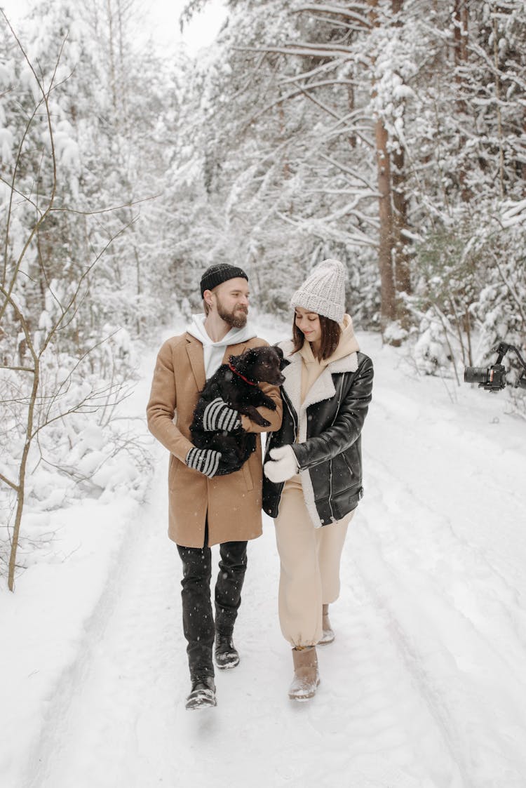 A Couple Walking On Snow Covered Ground Carrying A Dog