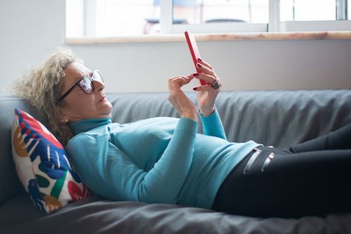 Free Elderly woman with glasses comfortably lies on a sofa indoors, using a smartphone. Stock Photo