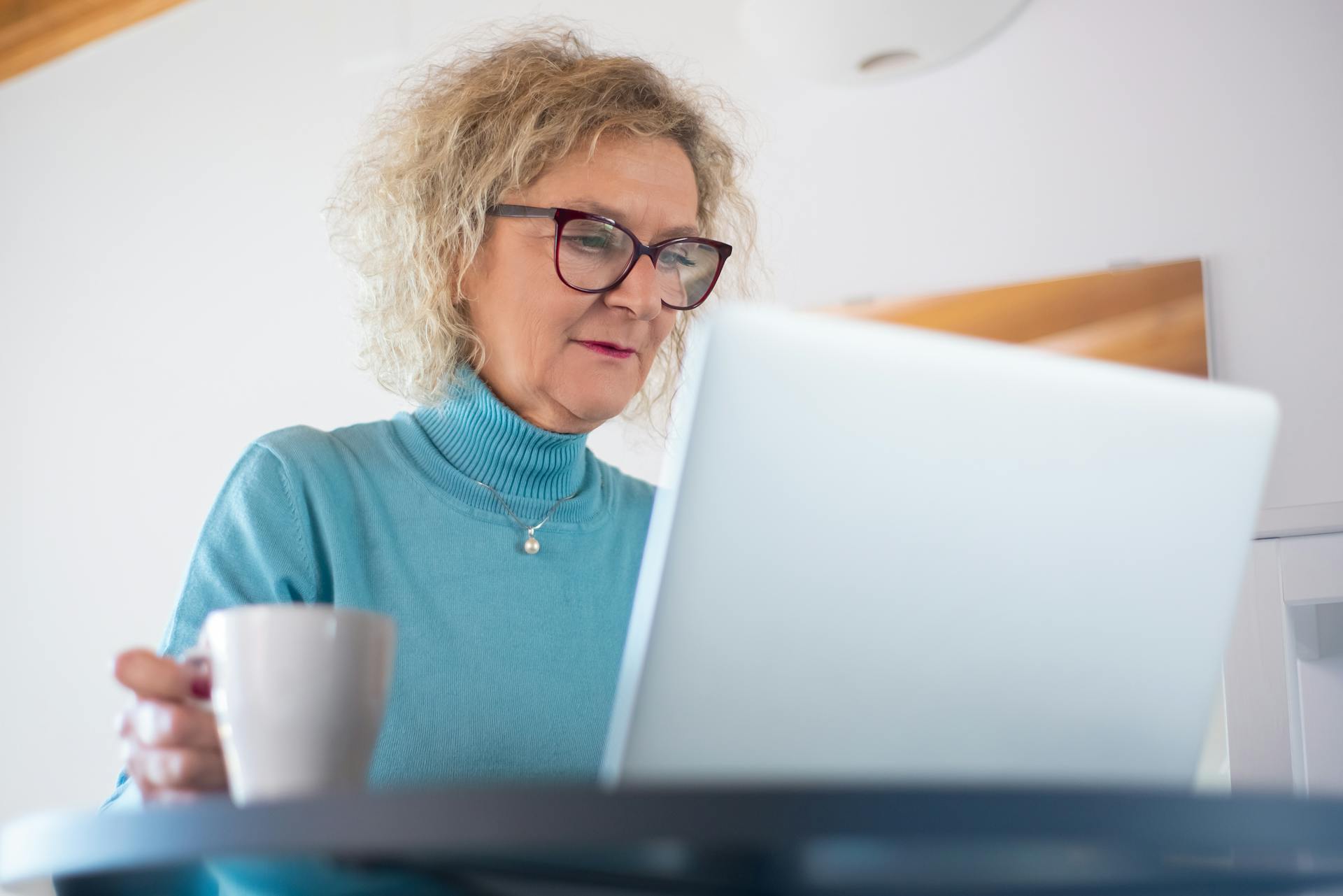 Elderly woman using a laptop at home with a cup of coffee, focusing on online work.