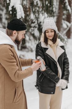 Couple in winter clothing sharing a warm drink from a thermos in a snowy forest.