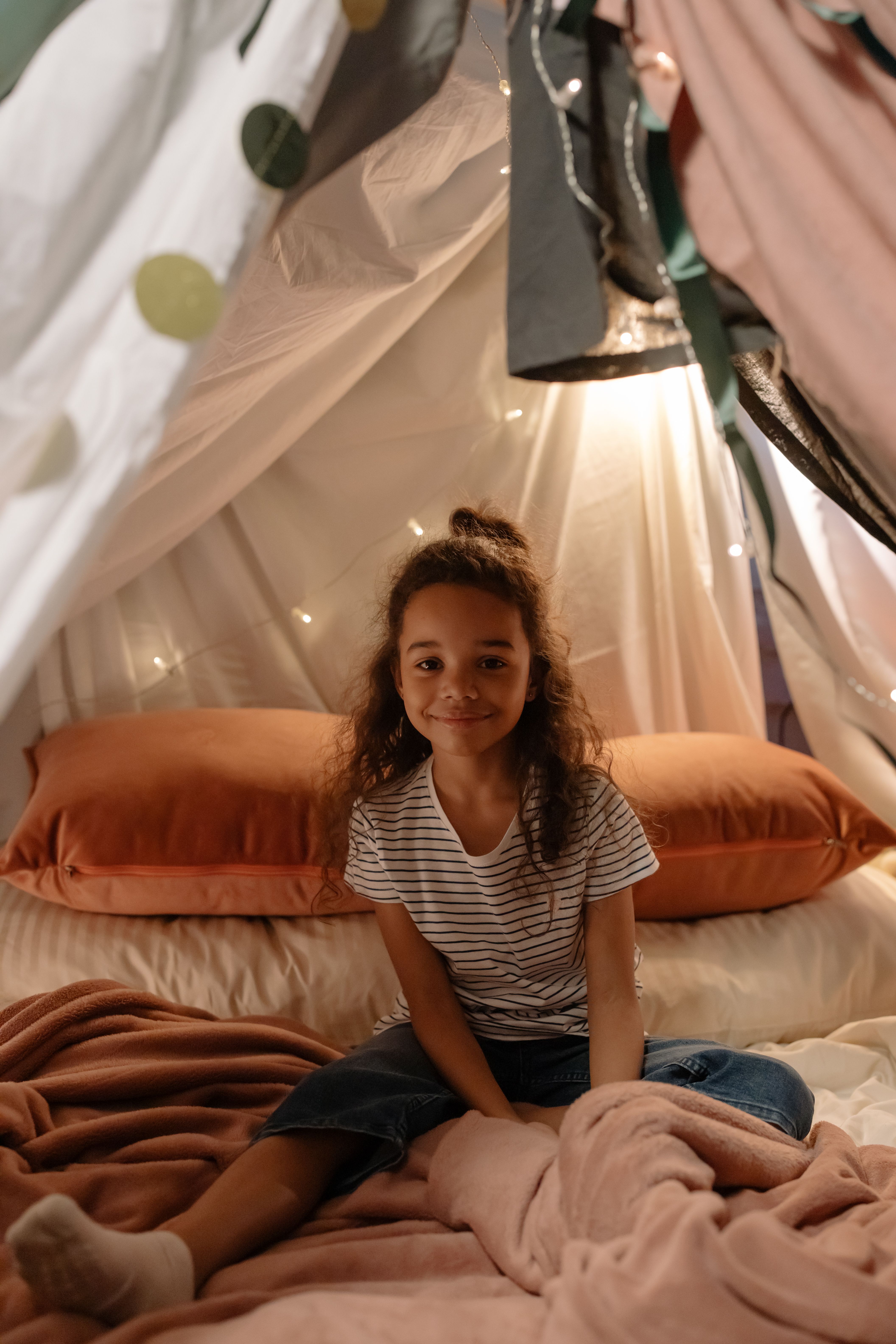 Cozy glamping setup with string lights, blankets, pillows, and a small tent in a backyard at dusk