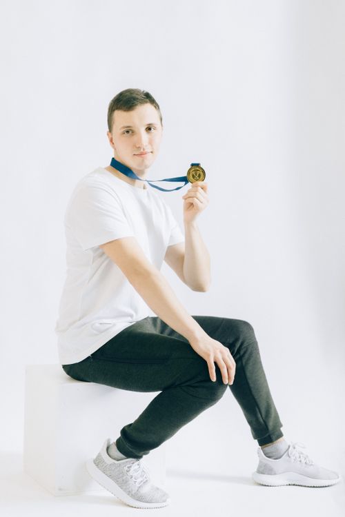 Free Smiling man in white t-shirt showcasing a gold medal while sitting on a stool in a studio. Stock Photo