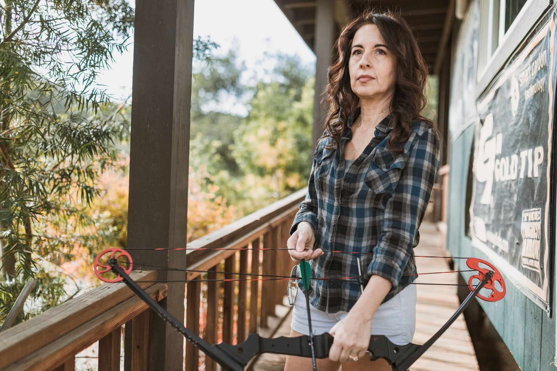 Woman holding a compound bow on a porch, ready to shoot outdoors.