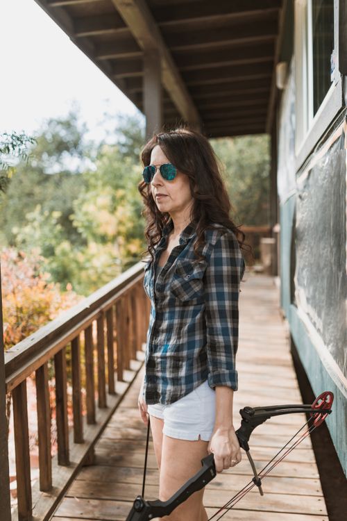 Free A woman with a bow stands on a wooden porch surrounded by nature, wearing sunglasses. Stock Photo
