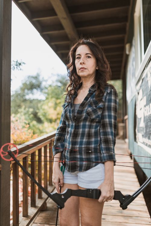 Free Woman holding bow and arrow on a wooden porch outdoors surrounded by nature. Stock Photo