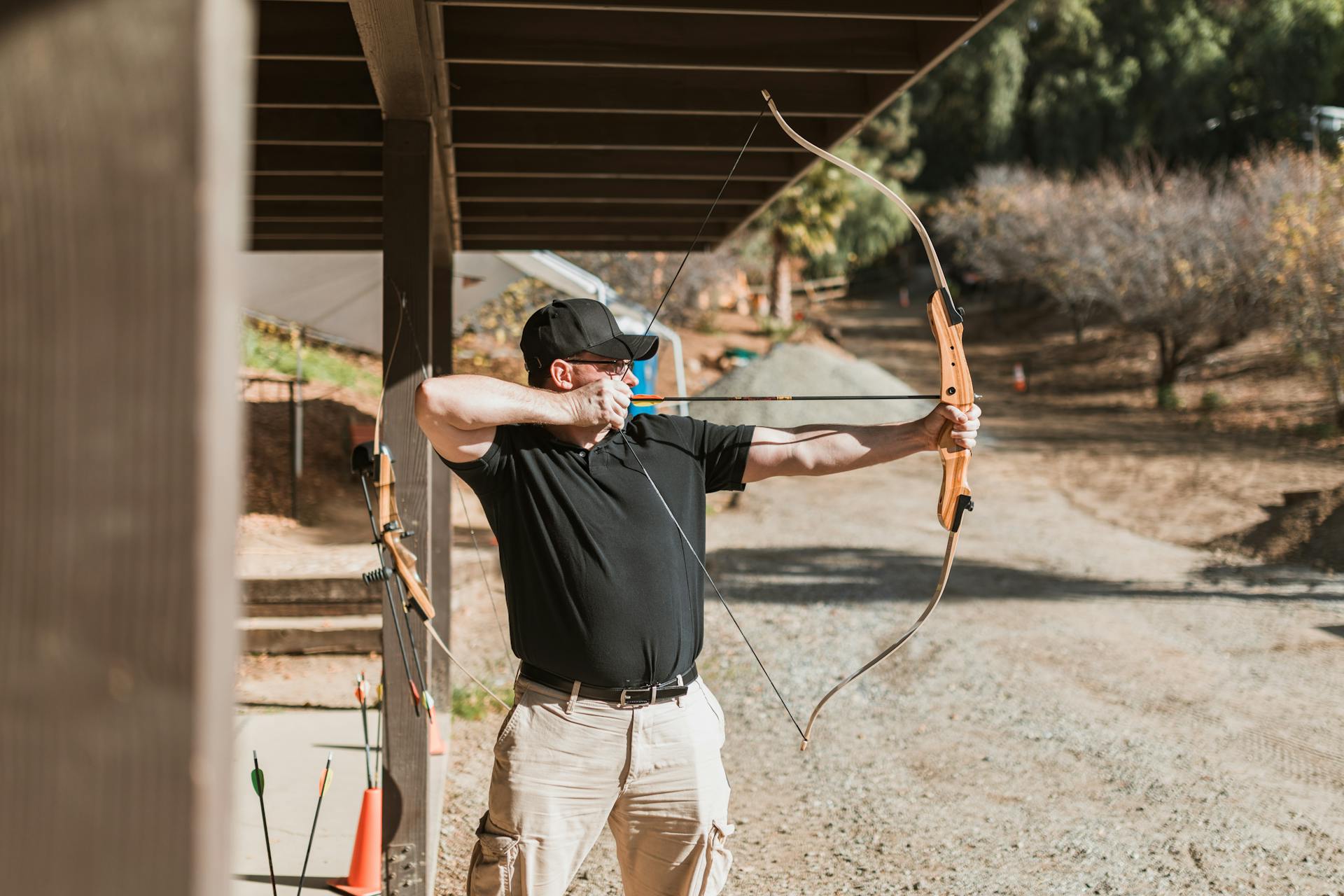 Adult male practicing archery outdoors. Wearing a black cap, focusing on target shooting. Sunny day.