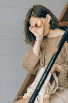 A woman sits on stairs, holding a net bag with a brown egg, playfully covering her face.