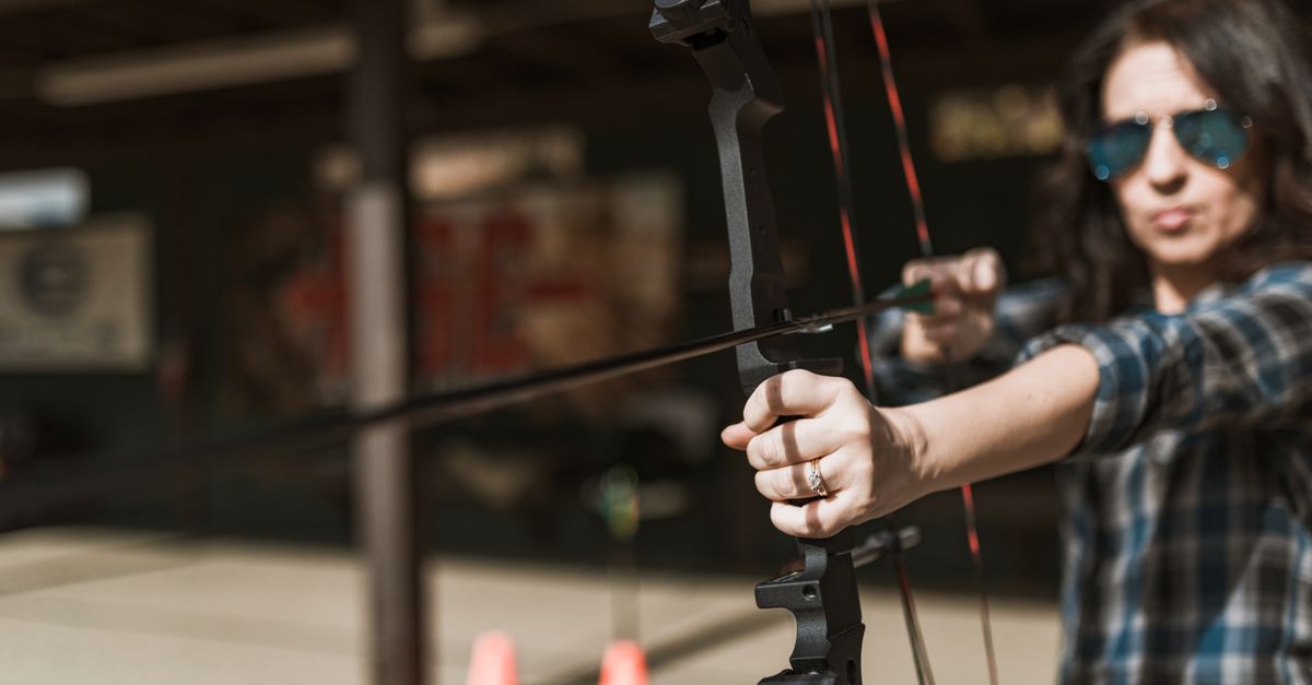 Woman with sunglasses aiming a compound bow outdoors, showcasing concentration and precision.