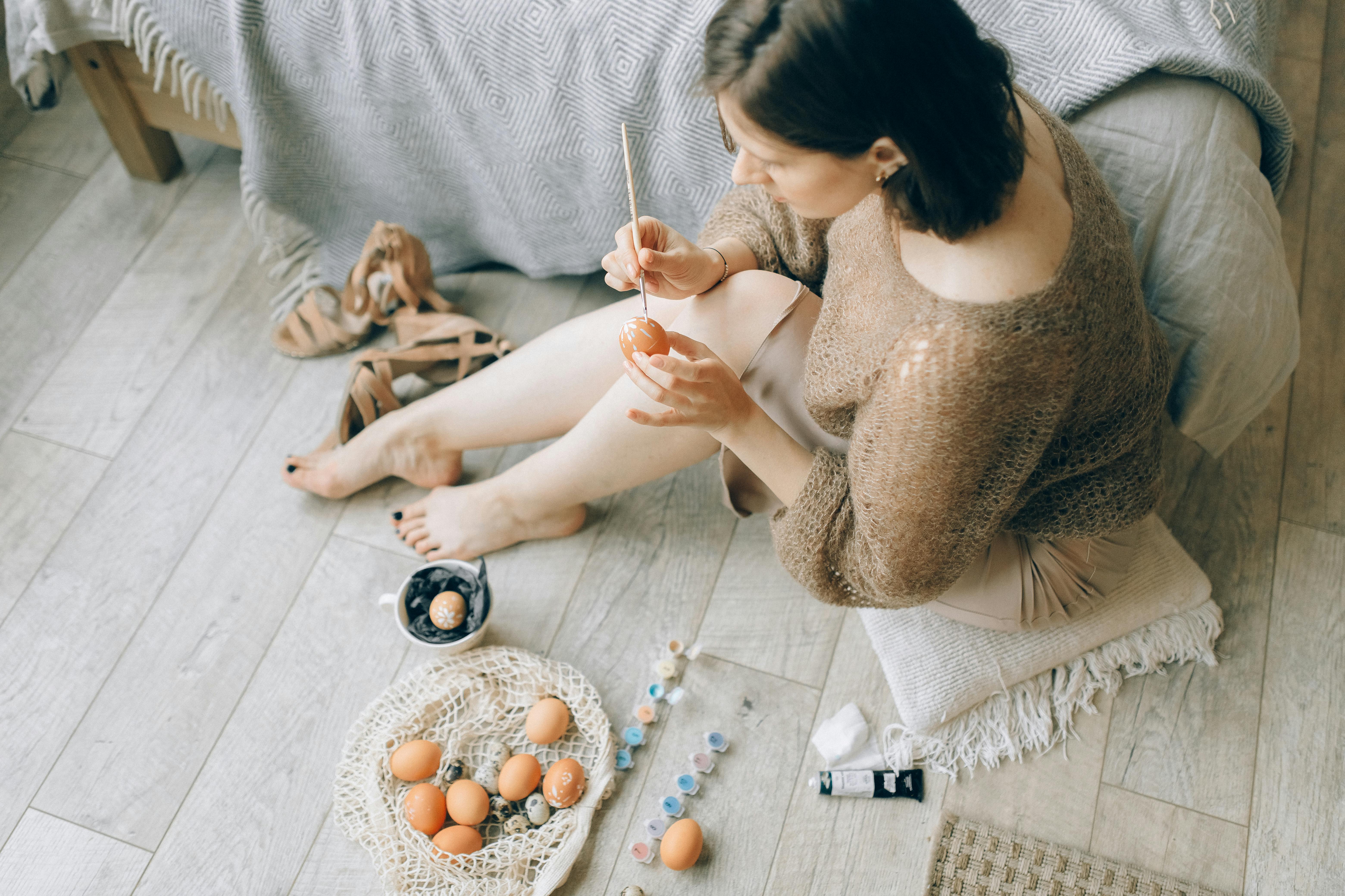 Woman sitting barefoot on wooden floor, painting Easter eggs, surrounded by art supplies.
