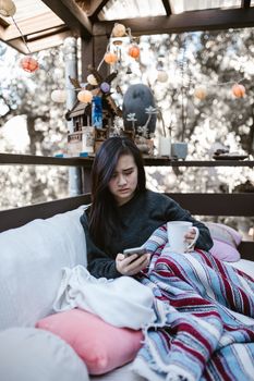 Woman wrapped in blanket, holding coffee on porch.