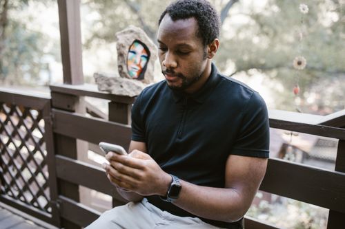 Free African American man sitting on a deck, focused on his smartphone outdoors. Stock Photo