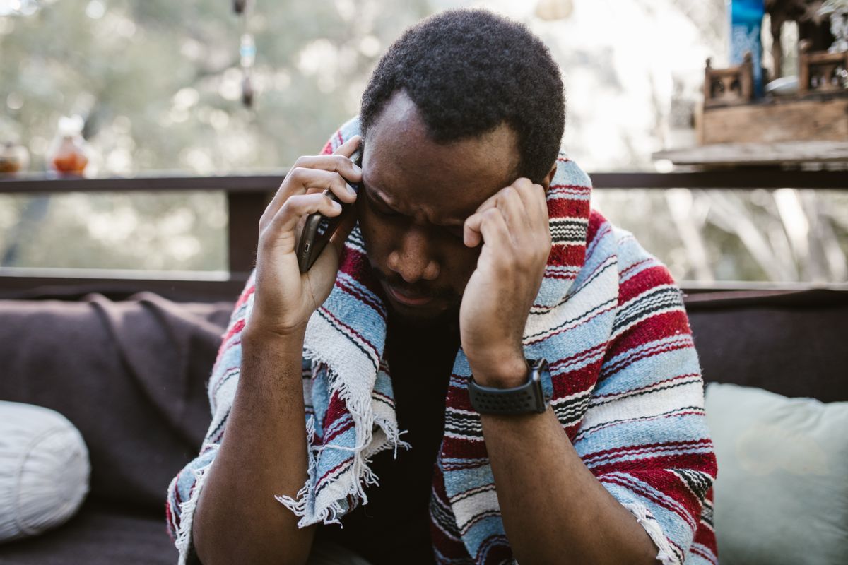A man sitting on a sofa wrapped in a striped red and blue blanket, holding a phone to one ear with his other hand pressed to his temple.