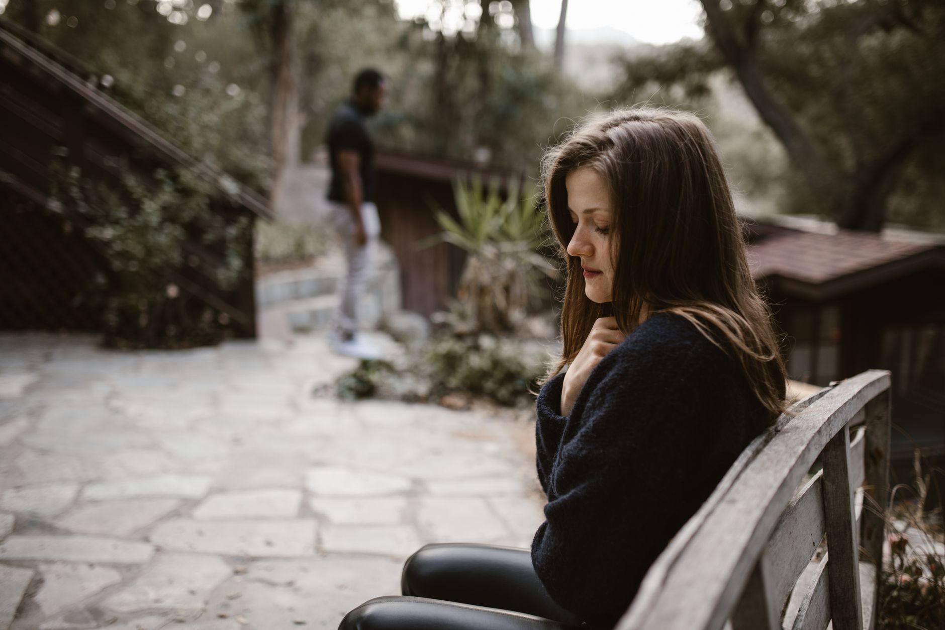 A woman sitting alone on a bench with a distant figure behind her, evoking a contemplative, cooled mood