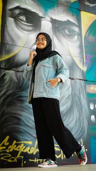 A smiling woman in a hijab poses confidently in front of colorful street art. - Fotografer Sidoarjo