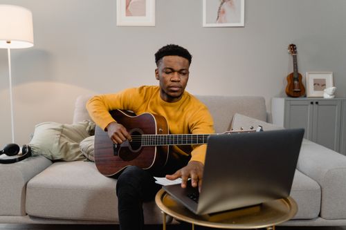 Free A man in a yellow sweater plays guitar while using a laptop in a cozy living room. Stock Photo