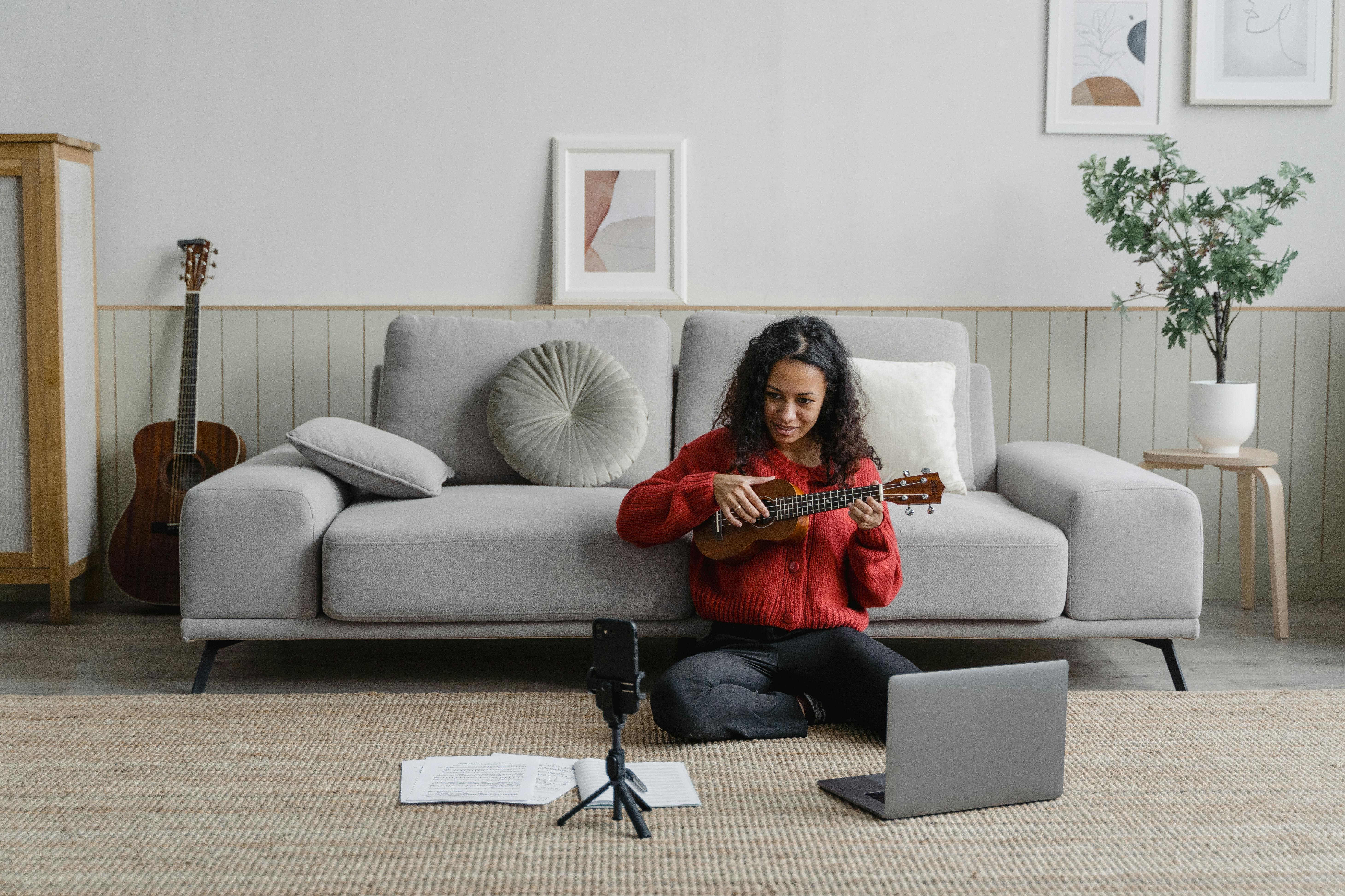 An adult woman sits on the floor playing a ukulele near a couch and laptop in a living room.