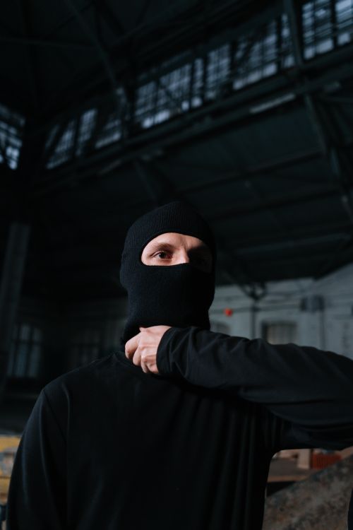 Free A person wearing a balaclava stands in a dimly lit industrial warehouse. Stock Photo