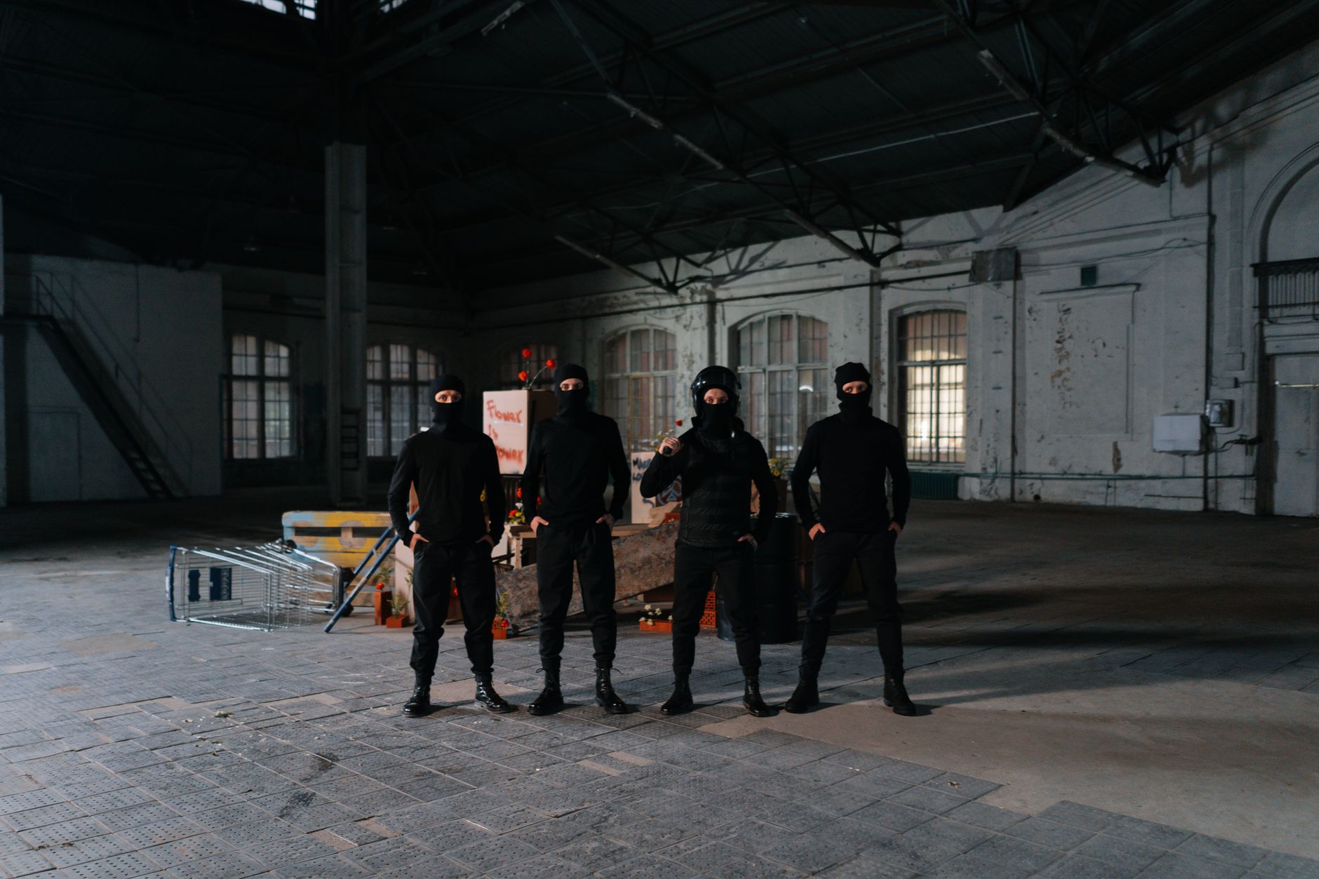 Four individuals in balaclavas pose in an empty warehouse setting, exuding a sense of secrecy.