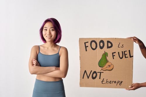 Free An Asian woman with purple hair smiles while holding a sign that reads 'Food is fuel, not therapy.' Stock Photo