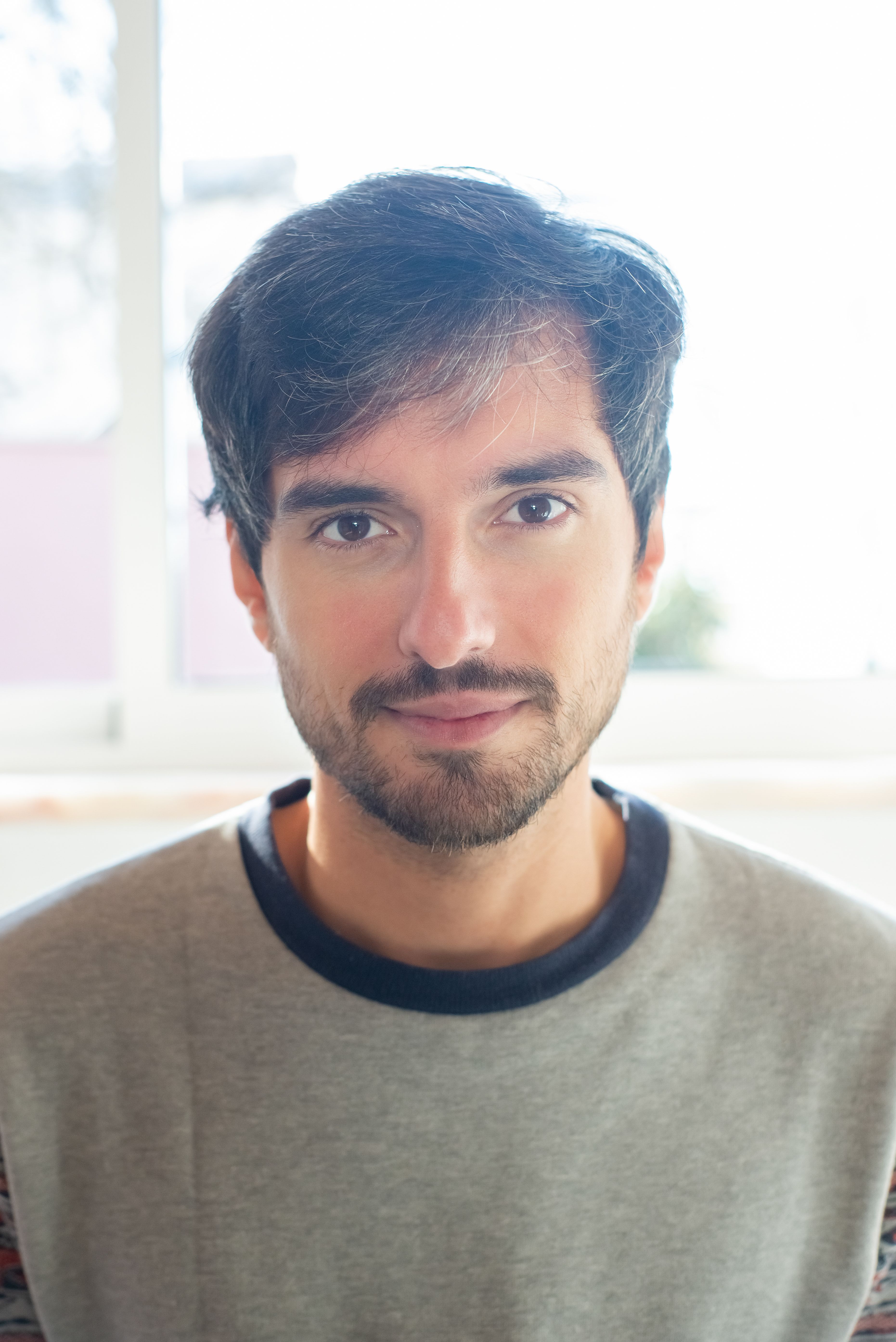 Close-up portrait of a bearded man wearing a gray shirt with a soft smile indoors.
