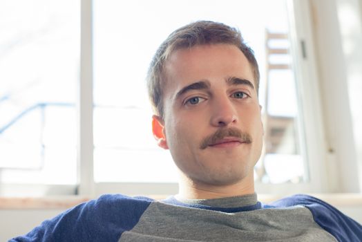 Portrait of a casually styled man with mustache relaxing indoors in natural light.