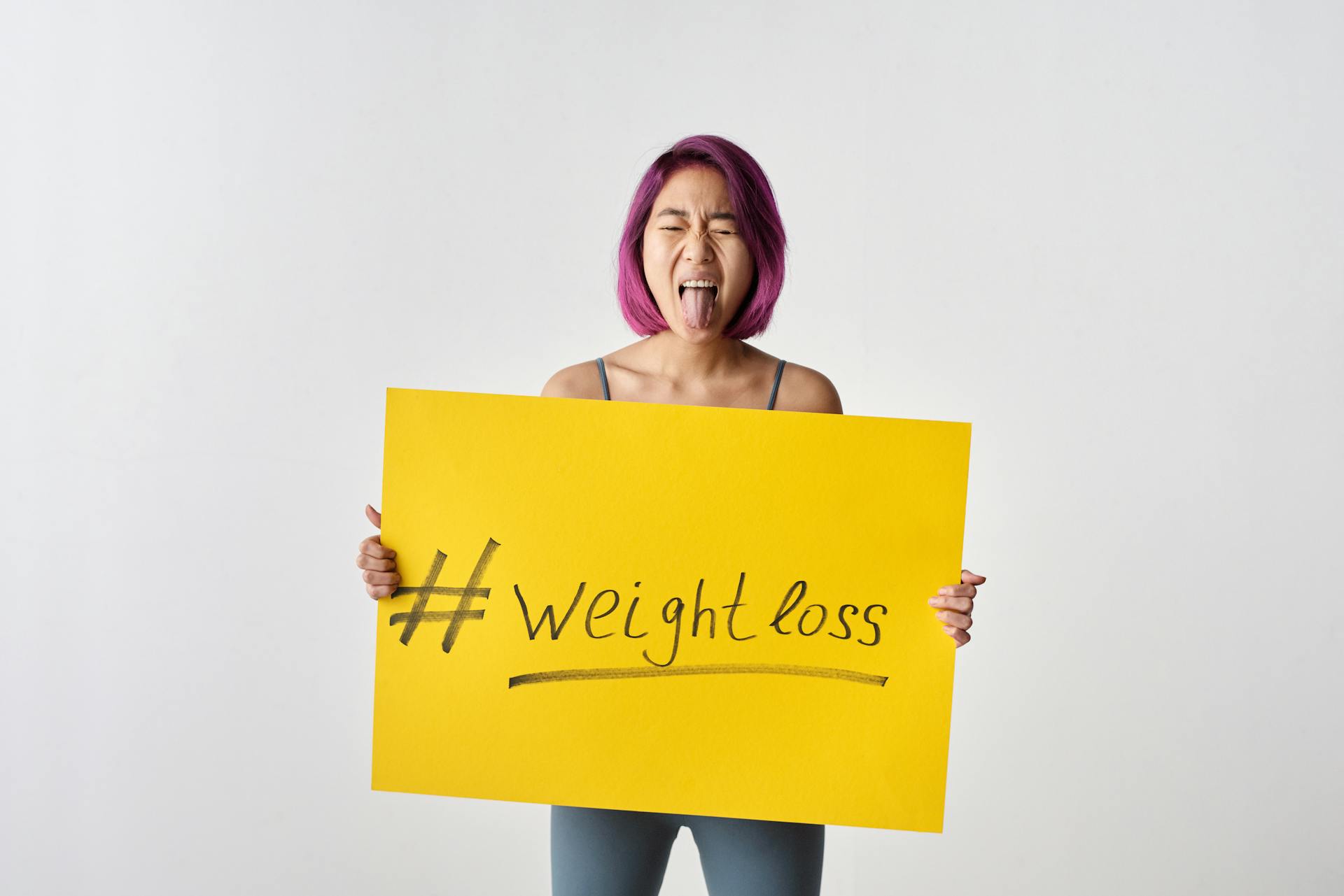 Young woman with purple hair holding a yellow weight loss poster in a studio shot.