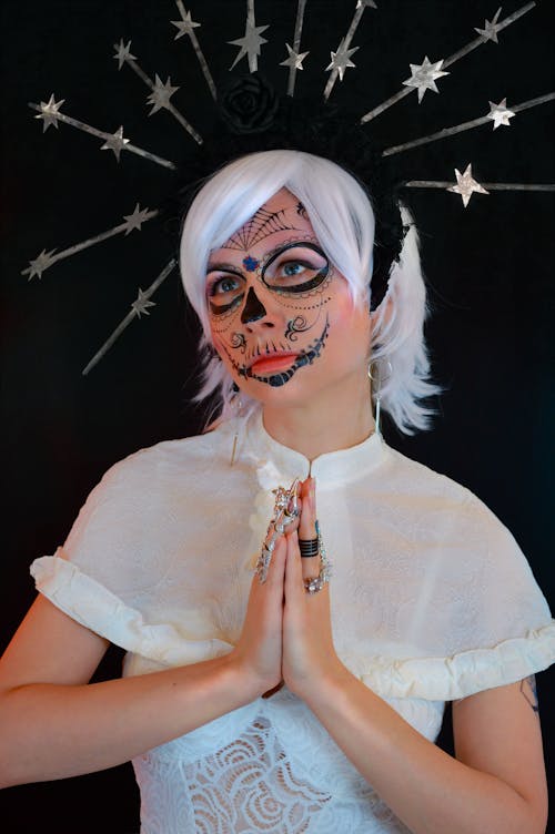 Serious young female magician with headband in white hair and creative makeup standing against black background with prayer hands