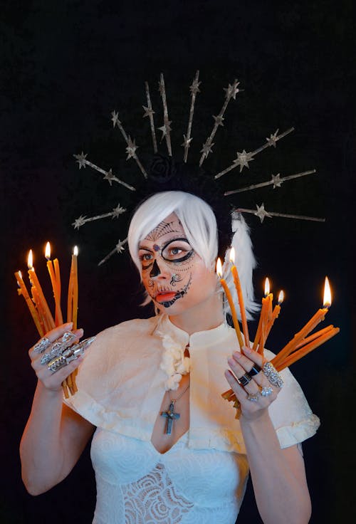 Serious young lady in white dress with creative headband and cross necklace holding burning candles in hands and looking away against black background during Mexican Day of the Dead