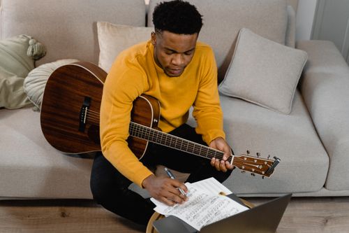 Free Young man with acoustic guitar studying music sheets indoors, learning online. Stock Photo