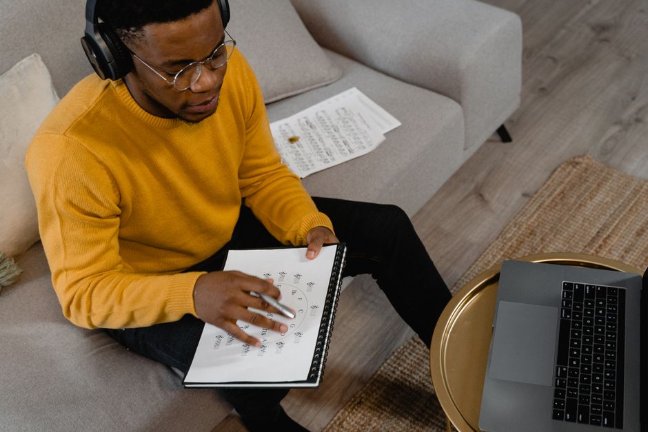 Man in mustard sweater using headphones, analyzing data with laptop and notebook indoors.