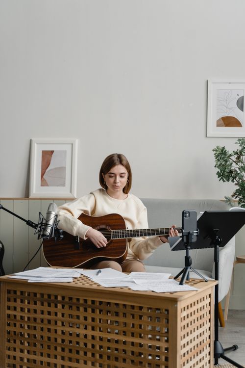 Free A woman playing an acoustic guitar in a cozy, modern living room setting with music sheets. Stock Photo