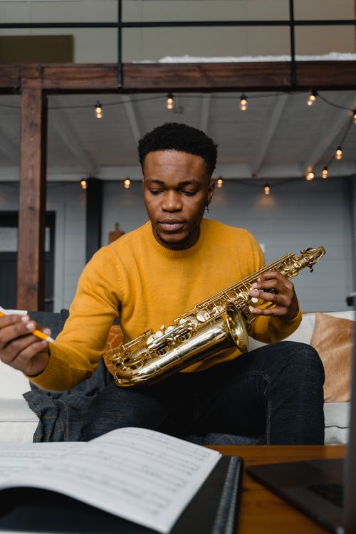 Free African American man in yellow sweater playing saxophone indoors with sheet music. Stock Photo