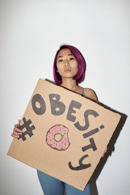 Free Asian woman with purple hair holding a hand-drawn obesity sign in a studio setting, promoting awareness. Stock Photo