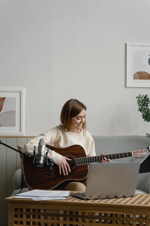 Free Caucasian woman playing guitar while smiling and sitting on a couch with a laptop nearby. Stock Photo