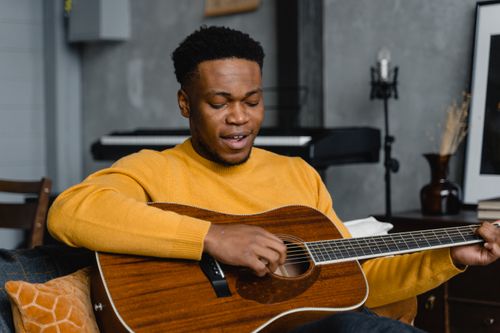 Free A young man wearing a yellow sweater plays an acoustic guitar indoors, creating a cozy musical ambiance. Stock Photo