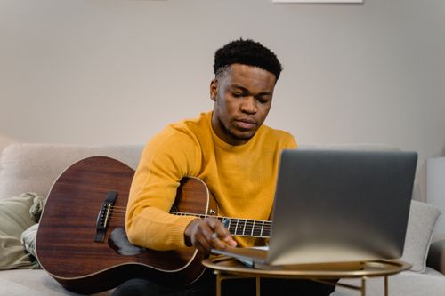 Free A man in a yellow sweater playing guitar and using a laptop to learn music indoors. Stock Photo