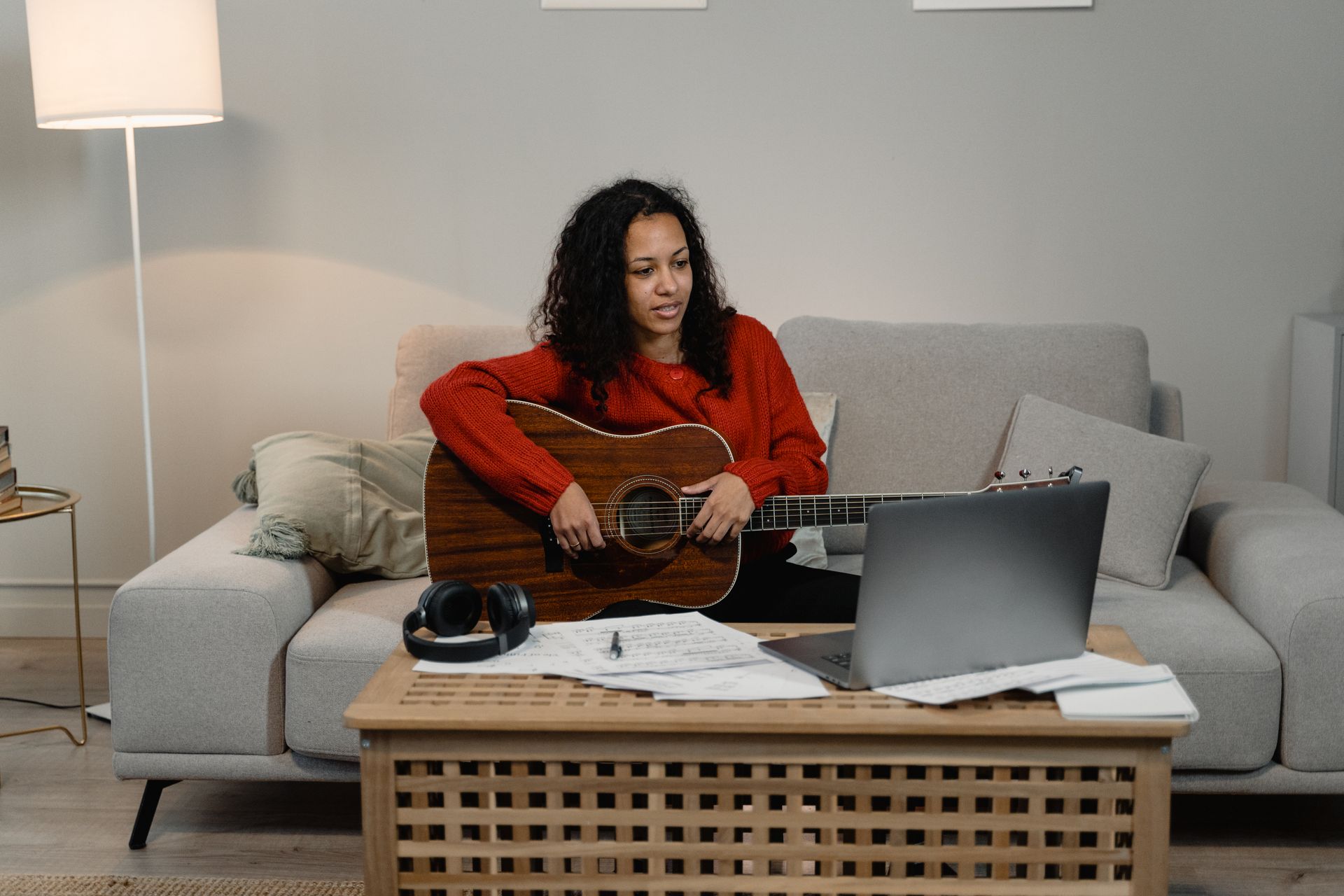 Woman in living room playing guitar while learning online via laptop. Cozy and creative setup.