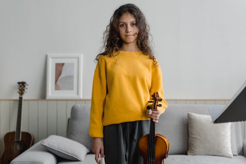 Free Teen girl holding violin in a cozy indoor setting, surrounded by musical instruments. Stock Photo