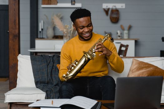 Cheerful man in a yellow sweater playing a saxophone on a couch in a cozy modern living room.